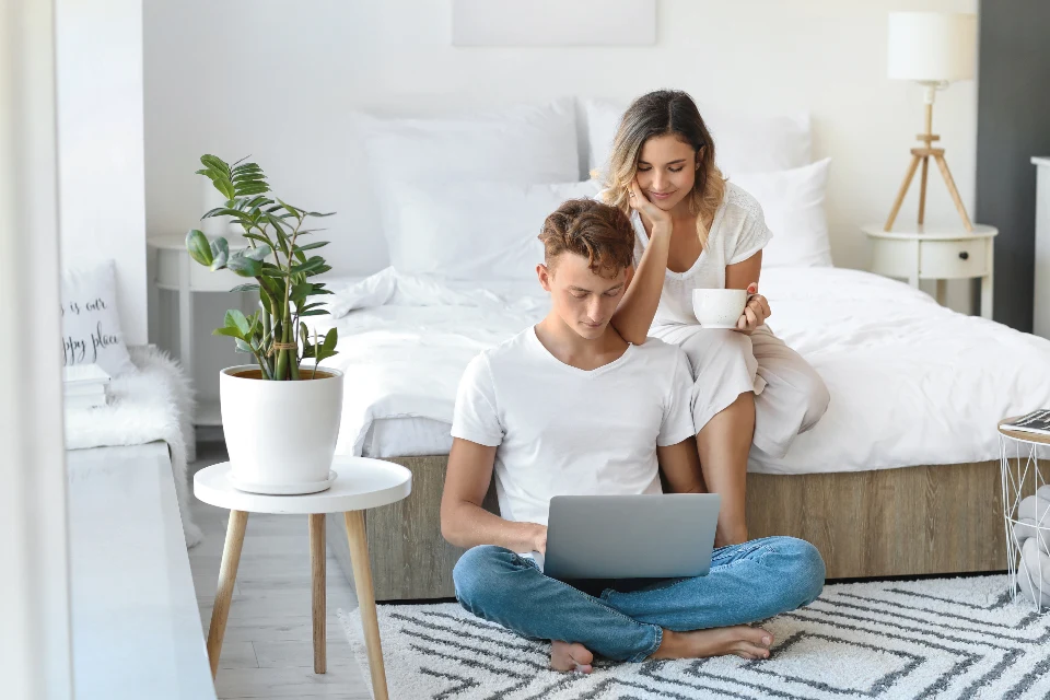 Pareja joven en casa: él sentado en el suelo con el portátil, ella detrás en la cama con una taza de café