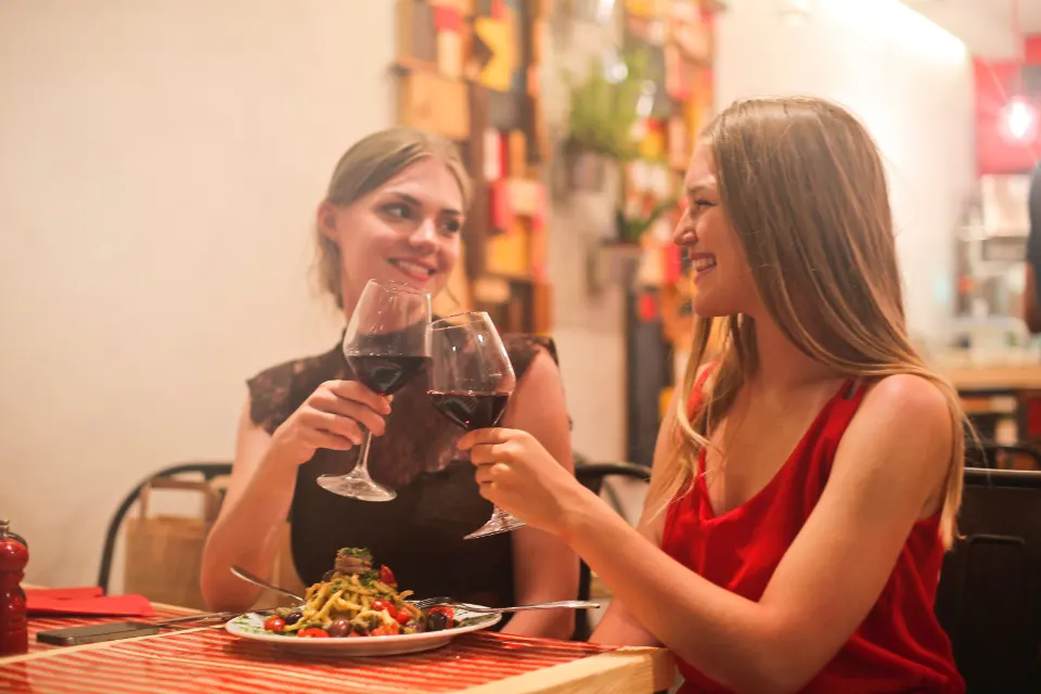 Zwei lächelnde Frauen stoßen mit Gläsern Rotwein in einem warm beleuchteten Restaurant an