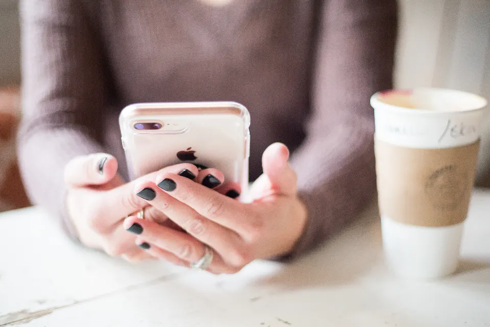 Hände einer Frau mit gepflegten Nägeln halten ein goldenes Smartphone über einem weißen Tisch, daneben ein Kaffee