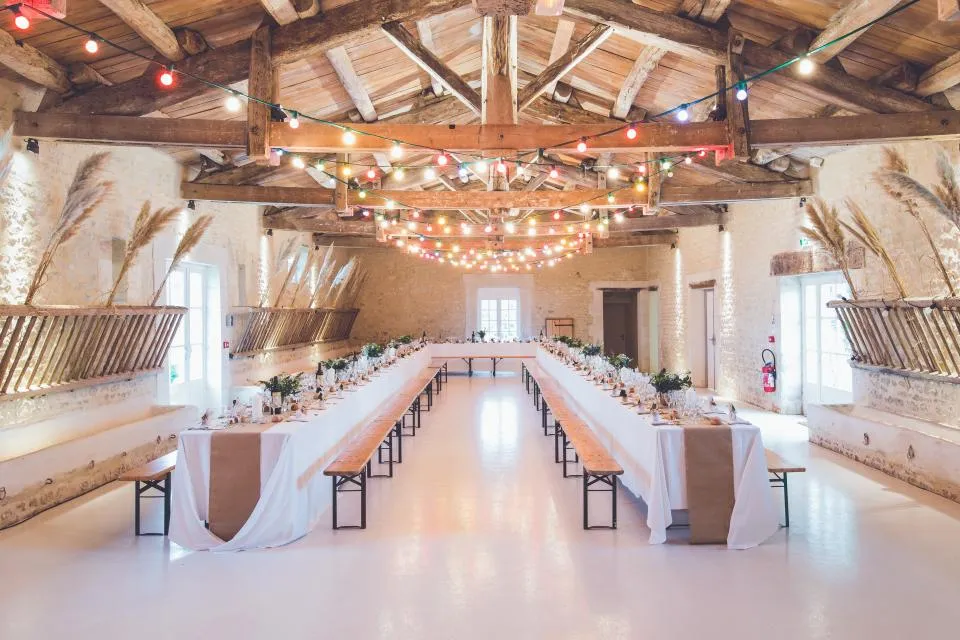 Salón rústico de celebraciones con vigas de madera, guirnaldas de luces y mesas largas vestidas de blanco